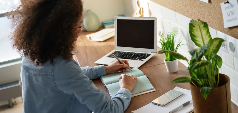 Mulher escrevendo em um caderno sobre a mesa, ao lado de um laptop, aplicando métodos de organização de estudos para a modalidade EaD.